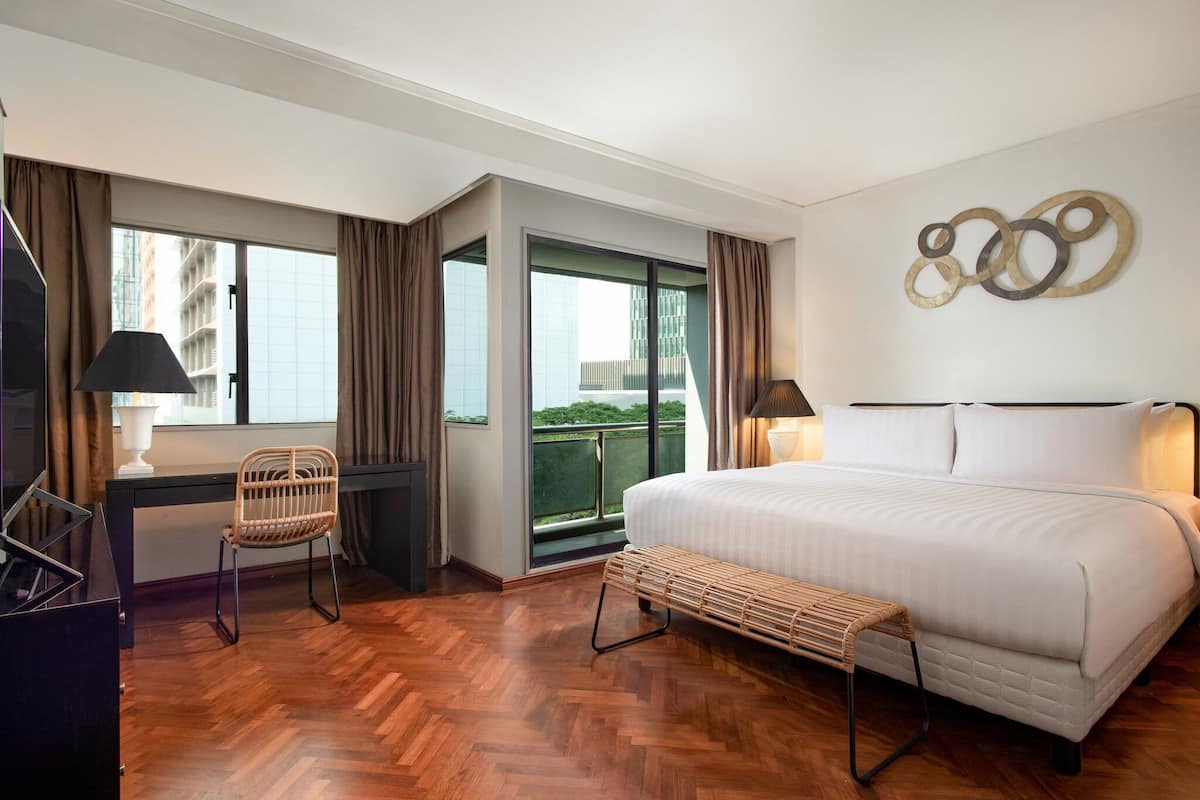 A hotel room with a large bed, a desk with a chair, a bench, and a view of a balcony.