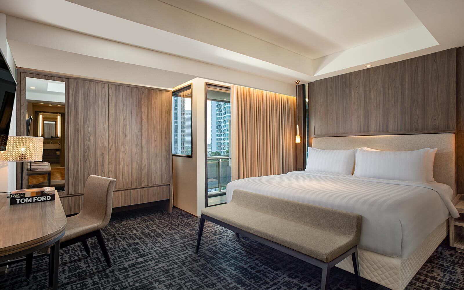 A hotel room with a large bed, a desk with a chair, a lamp, and a view of the city through the window.