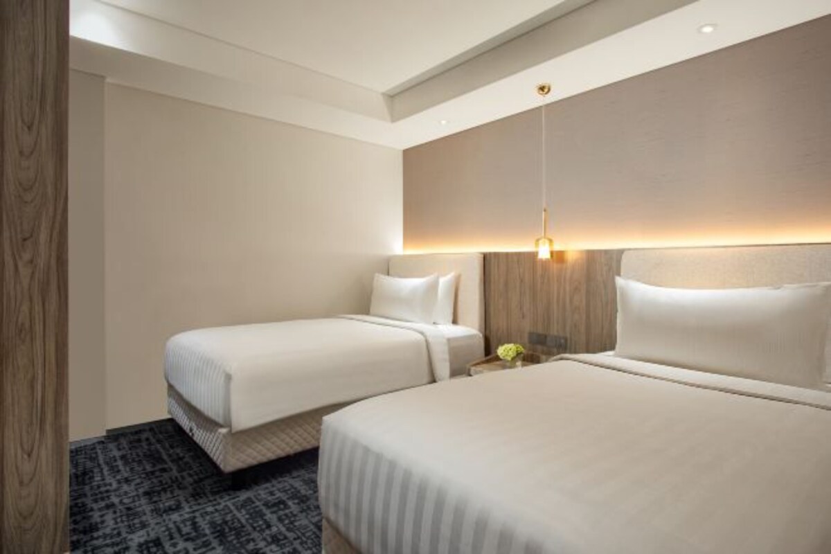 A hotel room with two beds, a wooden headboard, a small table with a lamp, and a patterned rug.