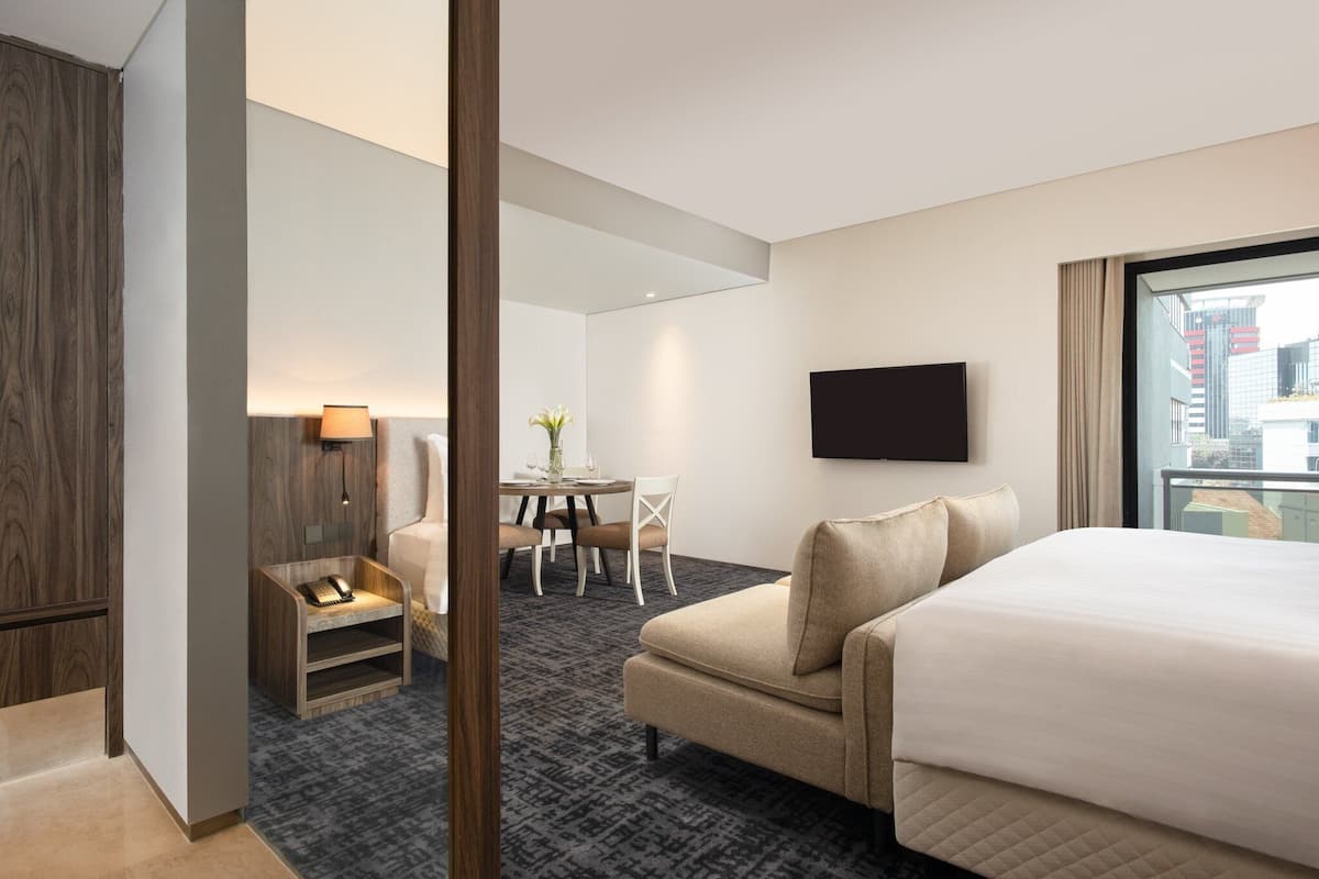 A hotel room with a large bed, a desk with a chair, a small table with a lamp, and a TV mounted on the wall.