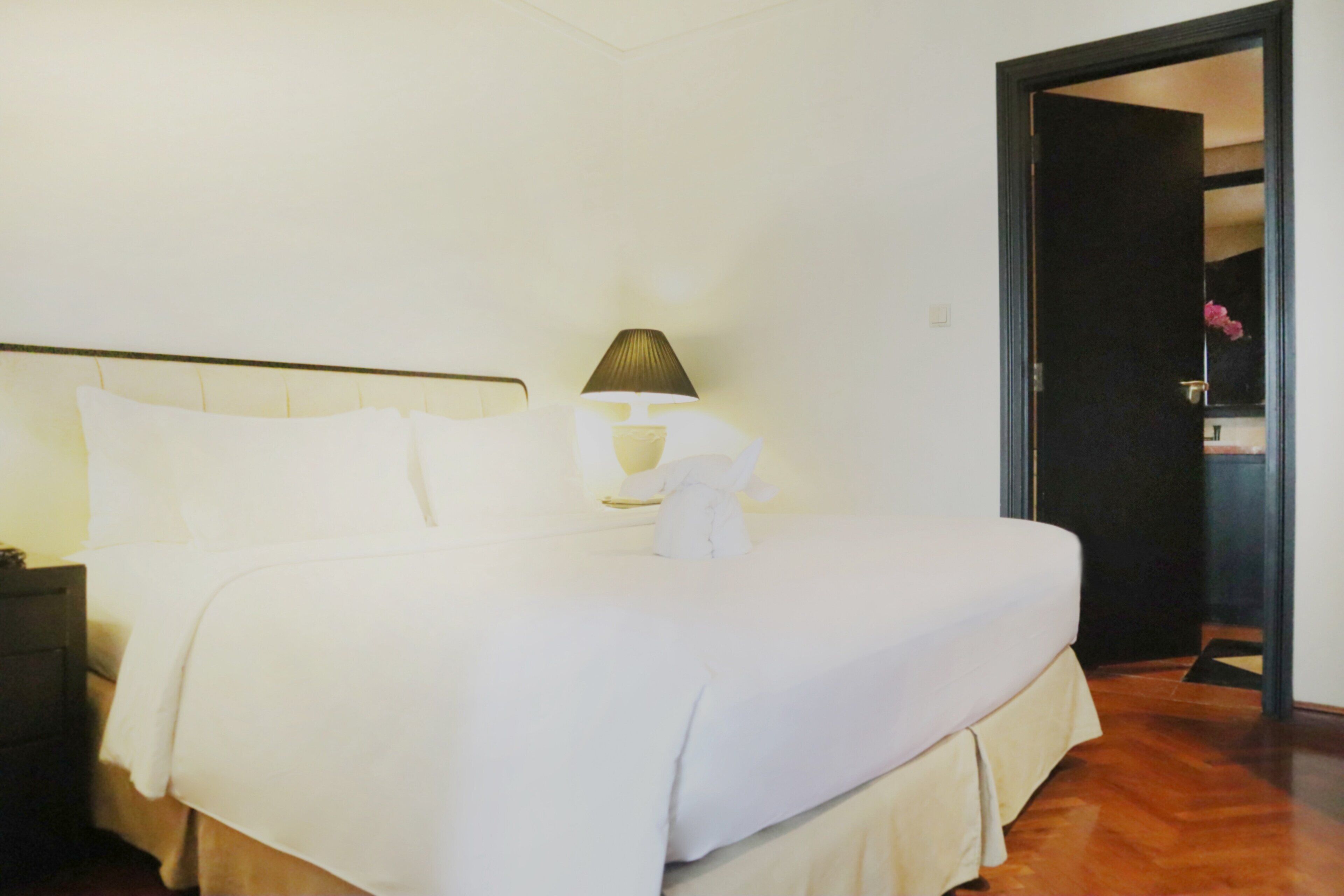 A neatly made bed with white linens, a bedside lamp, and a wooden floor.