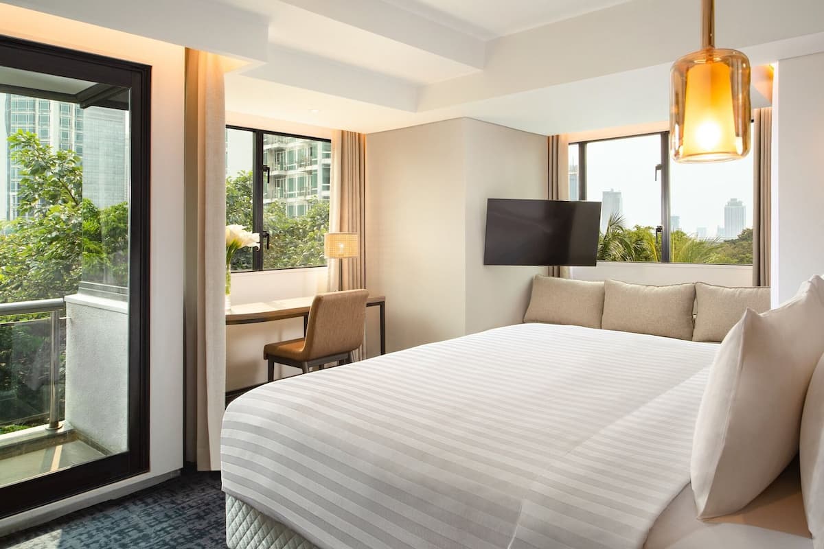A hotel room with a large bed, a desk, a chair, a TV, and a balcony with a city view.