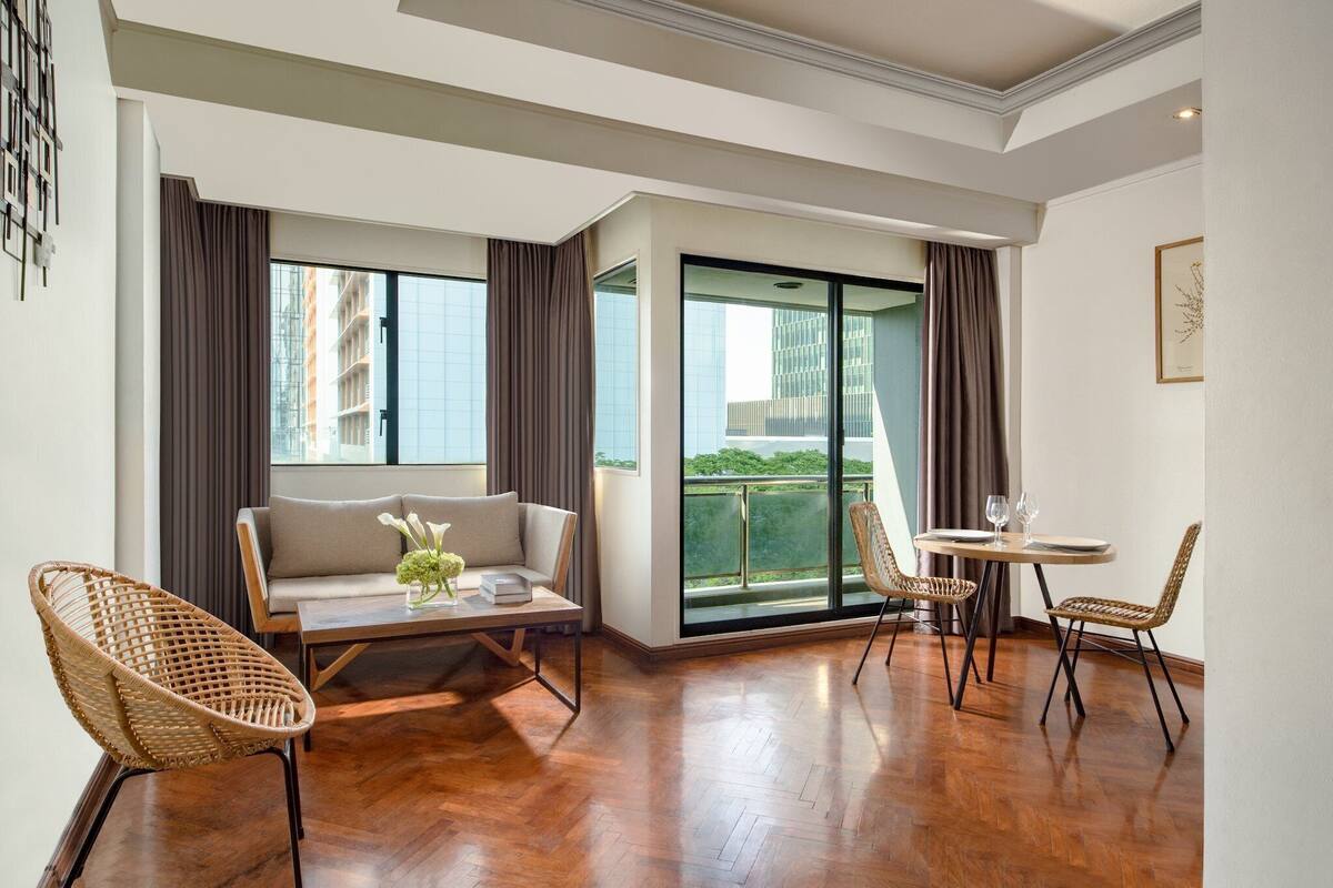 A modern living room with a sofa, two wicker chairs, a glass-top table, and a balcony with a view of buildings.