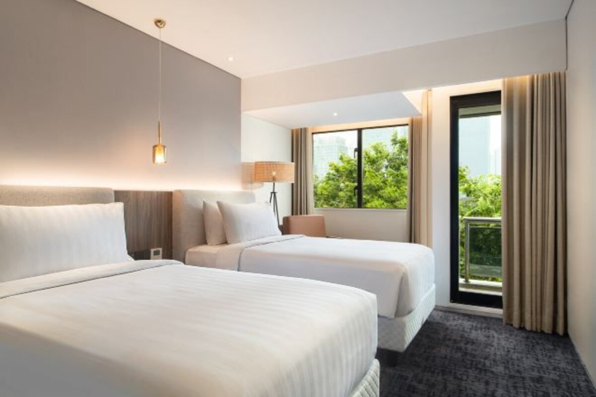 A hotel room with two beds, a large window with a view of greenery, a lamp, and a ceiling light.