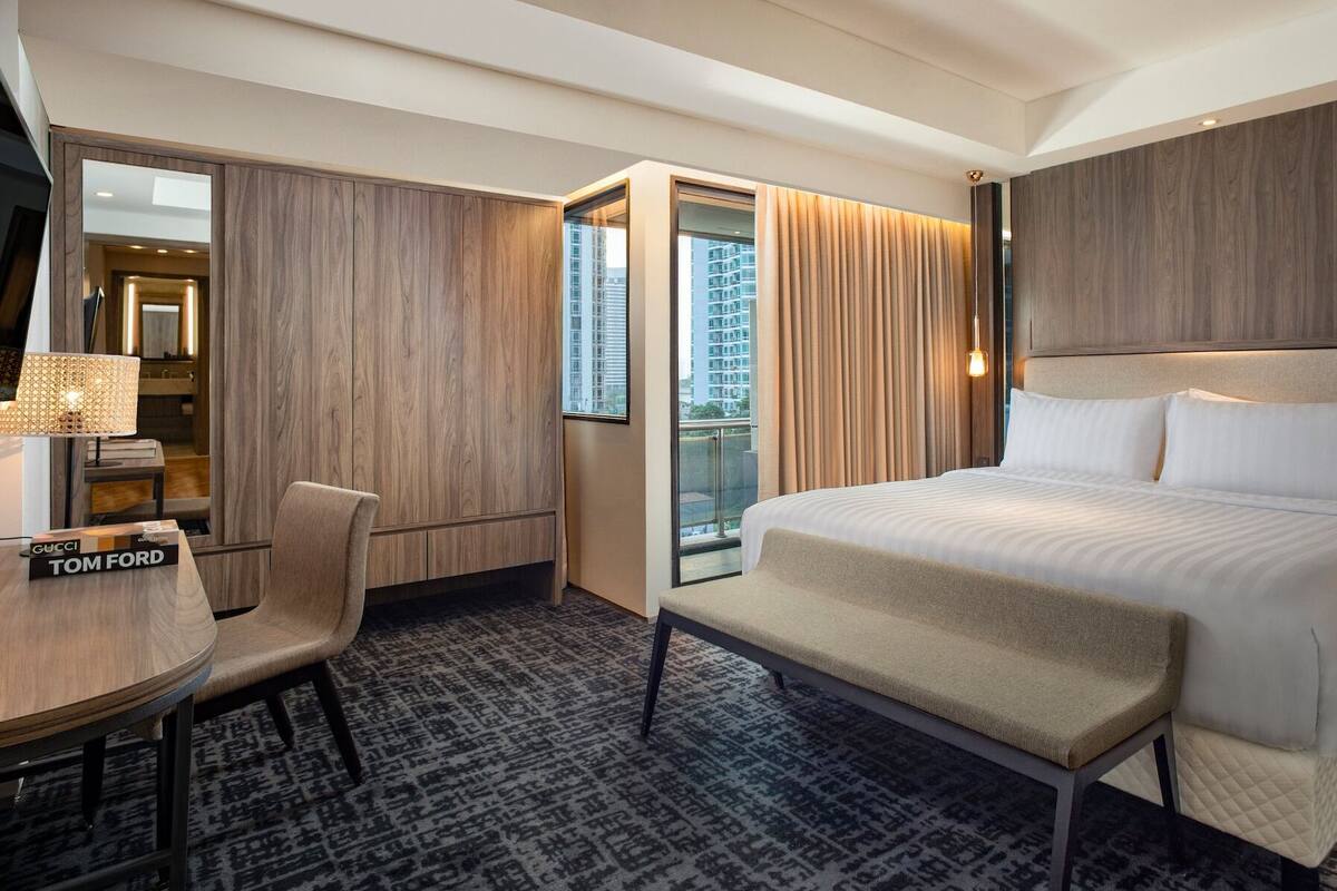A hotel room with a large bed, a desk with a chair, a wardrobe, and a view of the city through the window.