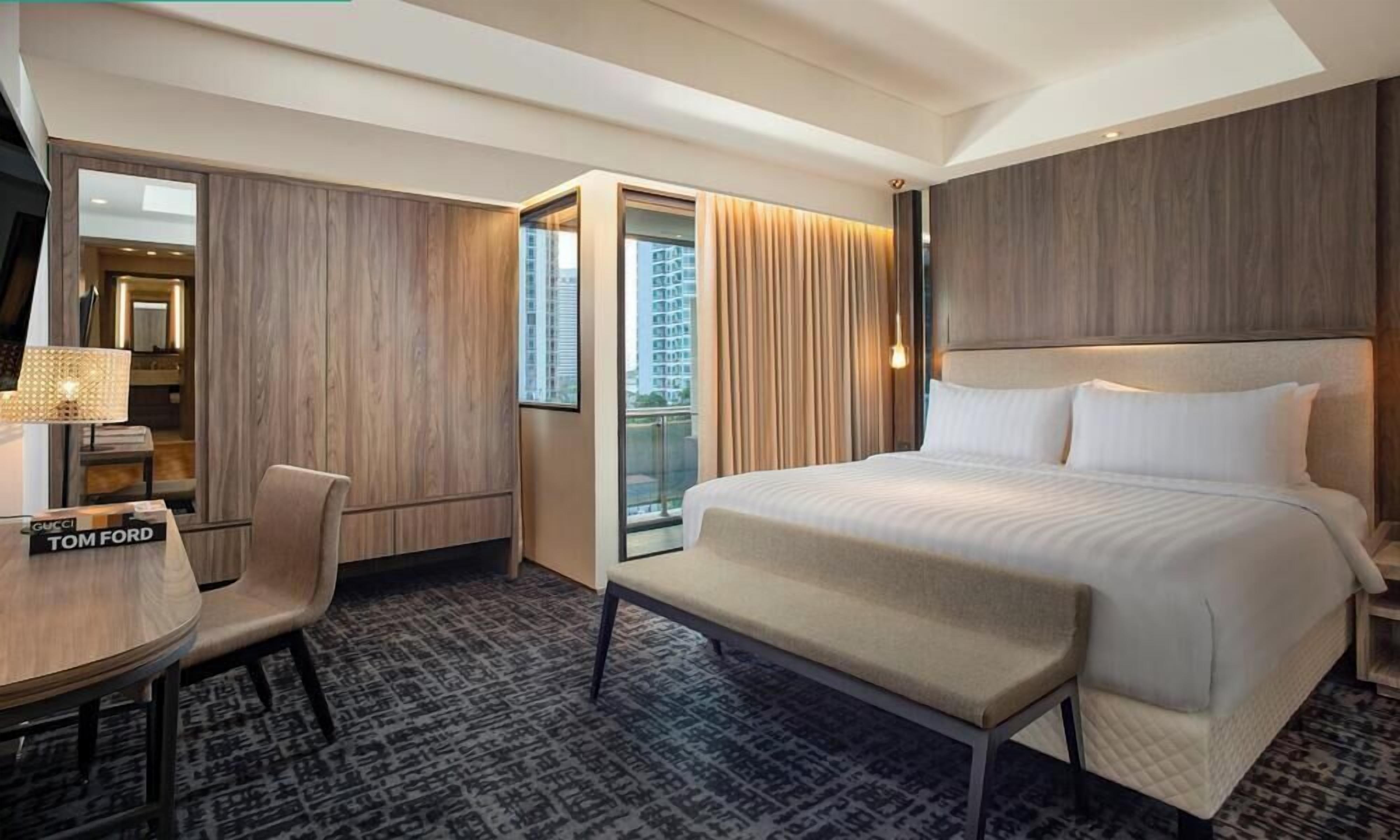 A hotel room with a large bed, a desk with a chair, a mirror, and a view of the city through the window.
