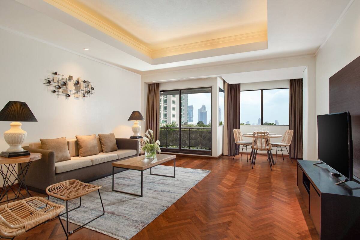 A modern living room with a sofa, coffee table, and TV stand. There is a balcony with a dining area and a view of the city.