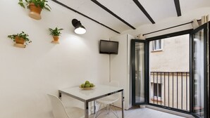 Una habitación pequeña y moderna con un ventanal de vidrio que da a un balcón, una mesa con sillas y plantas en macetas en la pared.