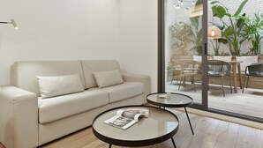 Ein modernes Wohnzimmer mit einer Couch, einem Glastisch und Blick auf eine Terrasse im Freien mit Pflanzen und Möbeln.
