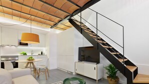 A modern kitchen with white cabinets, a dining area with a round table and chairs, a staircase with black railings, and a living area with a sofa and a green side table.