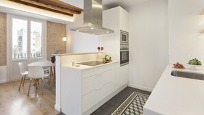 A modern kitchen with a built-in oven, a stainless steel range hood, and a dining area with a white table and chairs. - Comfortable and functional apartment in Plaza España (Barcelona)