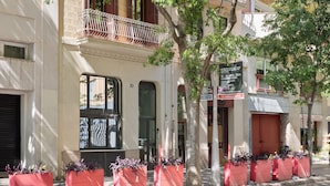 Une vue de rue montrant un immeuble doté d’un balcon, de grandes fenêtres et de jardinières rouges ornées de plantes.