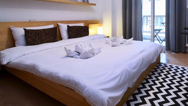 A hotel room with a large bed, a wooden headboard, a small shelf with decorative items, a view of the outdoors through a large window, and a black and white patterned rug. - New Apartment in the heart of Koukaki Orlof str (Athens)