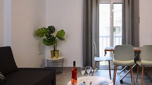 A modern living room with a sofa, a dining table with chairs, a potted plant, and a bottle of wine on the table. - New Apartment in the heart of Koukaki Orlof str (Athens)