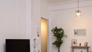 A modern living room with a flat-screen TV on a white stand, a potted plant, a small table with glasses and a bottle, and a mirror on the wall. - New Apartment in the heart of Koukaki Orlof str (Athens)