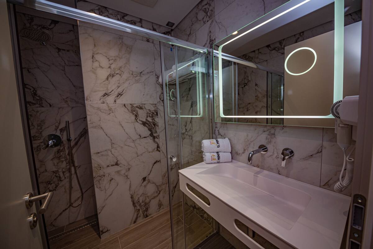 Ein modernes Badezimmer mit einer Glaskabine zur Dusche, einem weißen Waschbecken und einem großen Spiegel mit runder Beleuchtung.