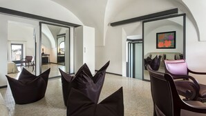 Amplia habitación con techos altos, un ventanal y vistas a la ciudad. Hay sillas modernas y un sofá con un cojín decorativo.