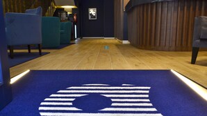 Vestíbulo de hotel con alfombra azul que presenta un logotipo, suelo de madera, sillas color turquesa y un mostrador de recepción de madera.