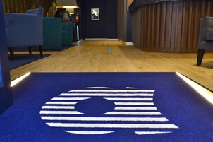 A hotel lobby with a blue carpet featuring a logo, wooden flooring, teal chairs, and a wooden reception desk.