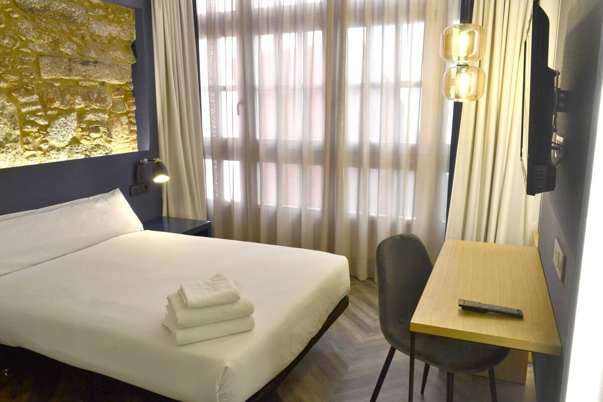 Quarto de hotel com cama, escrivaninha, cadeira, controle remoto e luminária na parede.