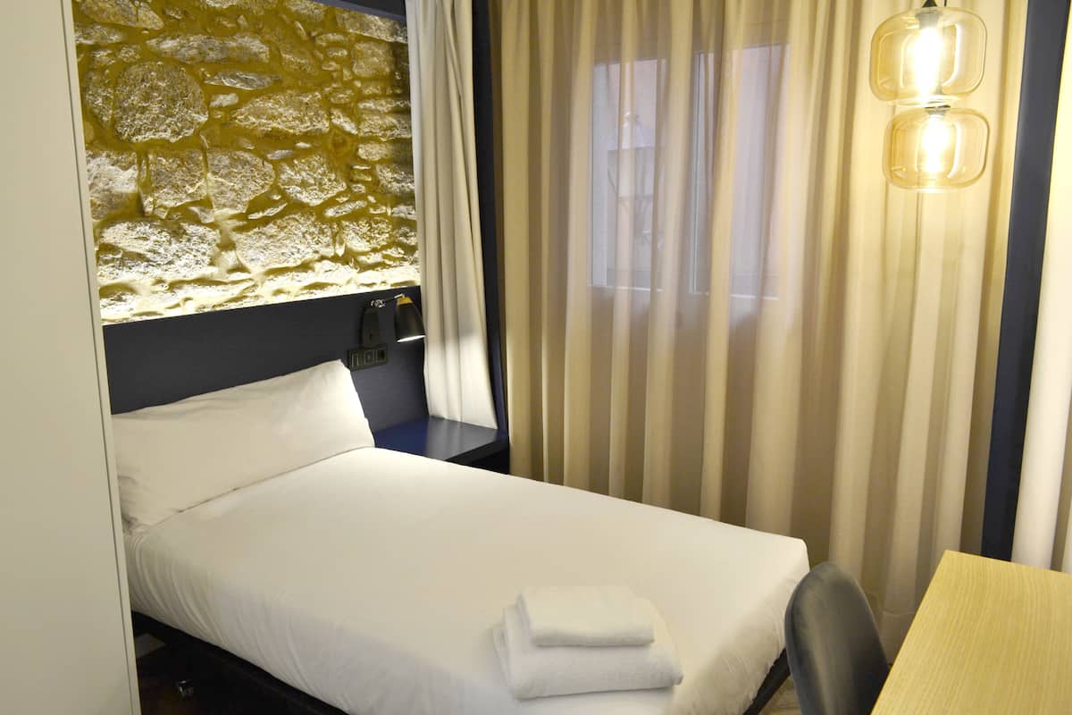 Quarto de hotel com cama, escrivaninha, luminária e janela com cortinas.
