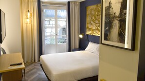 Habitación de hotel con cama, escritorio, silla, ventana con cortinas, lámpara de pared y un cuadro enmarcado.