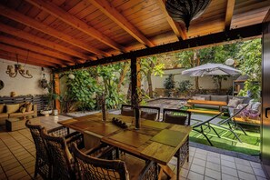 Un patio al aire libre con mesa de madera y sillas de mimbre, rodeado de vegetación y un muro de ladrillo.