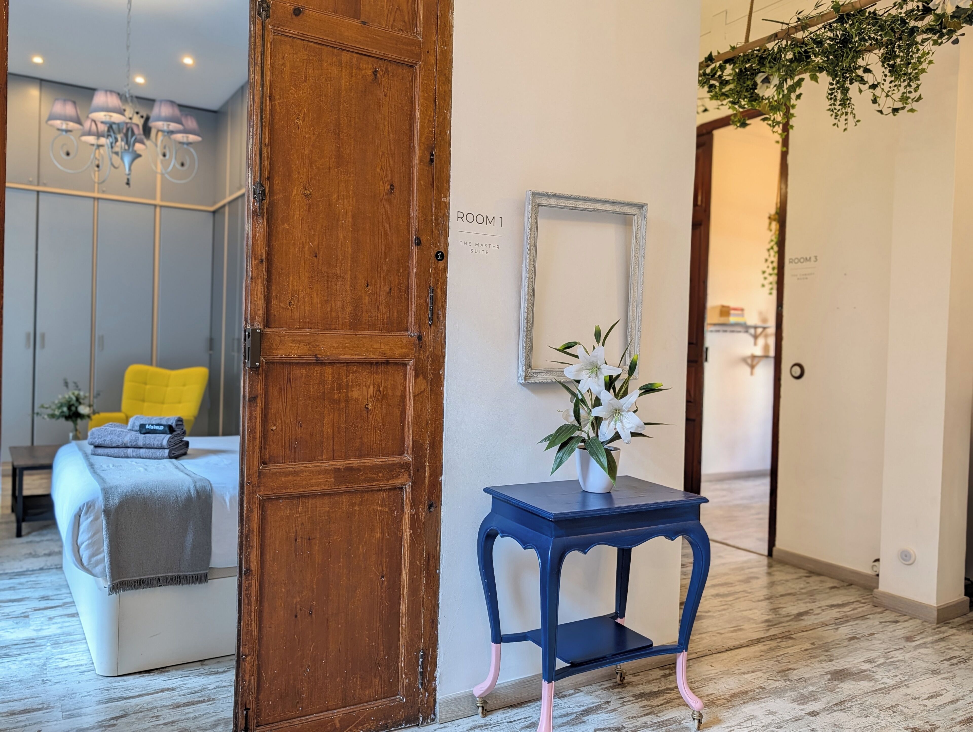 A hotel room with a wooden door, a bed with a grey blanket, a small table with a vase of flowers, and a mirror on the wall.
