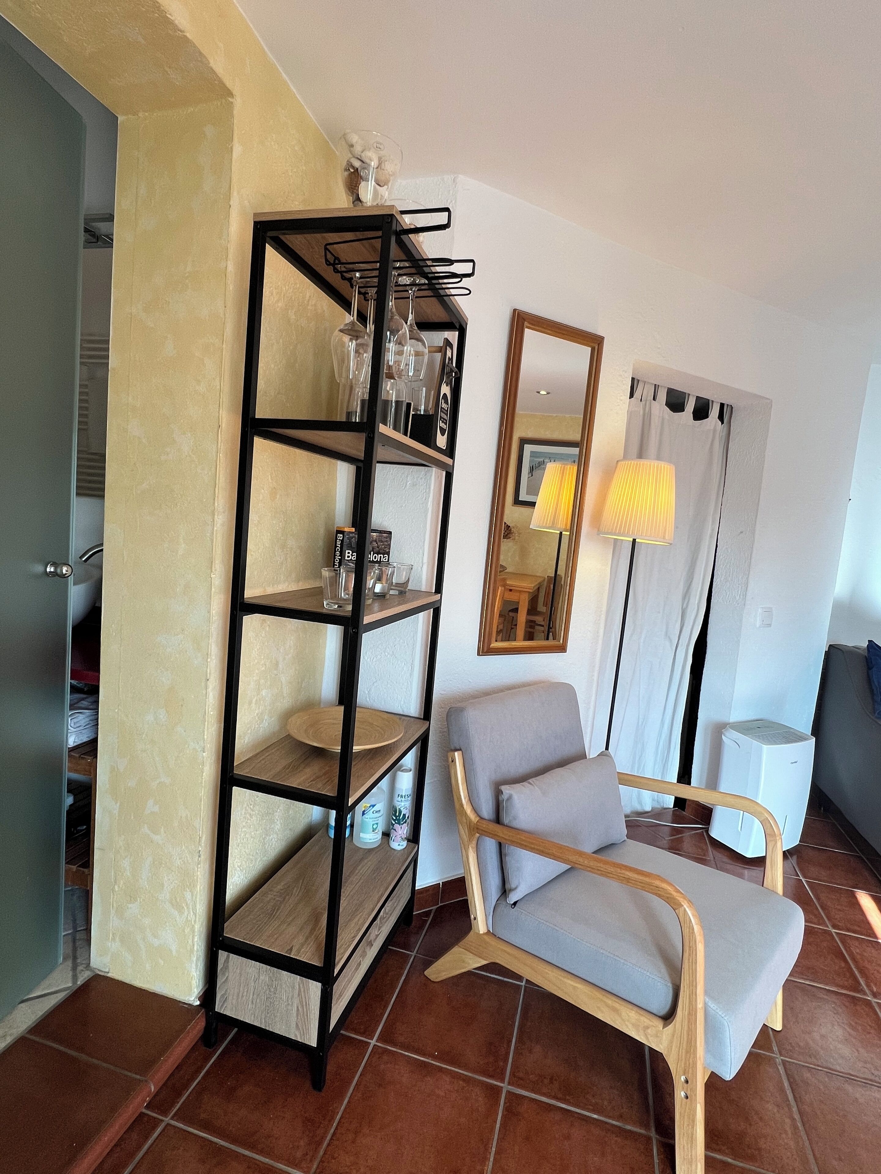 Una sala de estar con una silla de madera, un estante de metal negro con cristalería, una lámpara y un purificador de aire blanco.