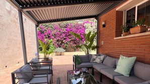 An outdoor patio with a sofa, chairs, and a table. There are potted plants and a view of a garden with pink flowers. - Beautiful apartment, garden and swimming pool, 20km from Barcelona, suitable for pets (Vallirana)