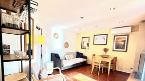A living room with a sofa, a dining table with chairs, a wooden chair, a TV, and a shelf with glasses and a lamp. - Beautiful apartment, garden and swimming pool, 20km from Barcelona, suitable for pets (Vallirana)