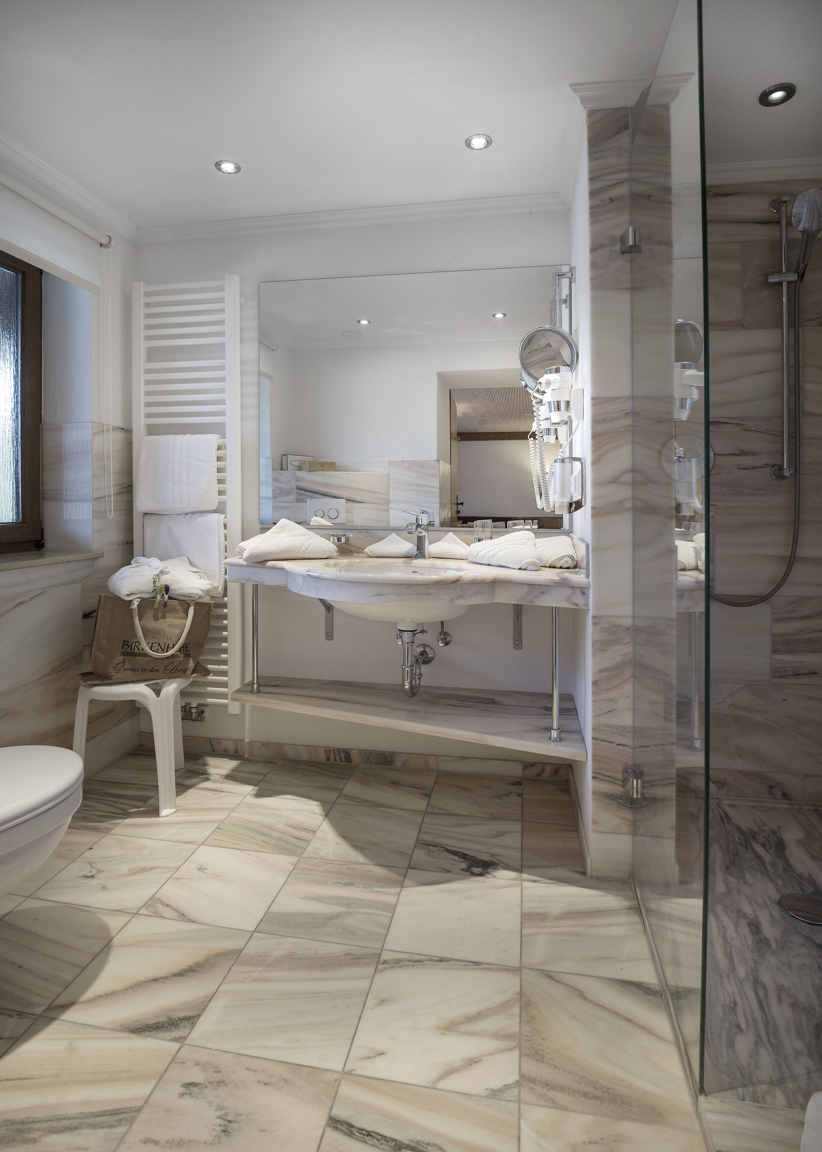 A modern bathroom with a marble floor, a large mirror, a glass shower enclosure, and a white vanity with a towel rack.