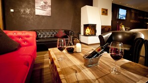 A cozy living room with a fireplace, a red sofa, a wooden table with wine glasses and bottles, and a television on the wall. - Hotel Birkenhöhe (Mittelberg)