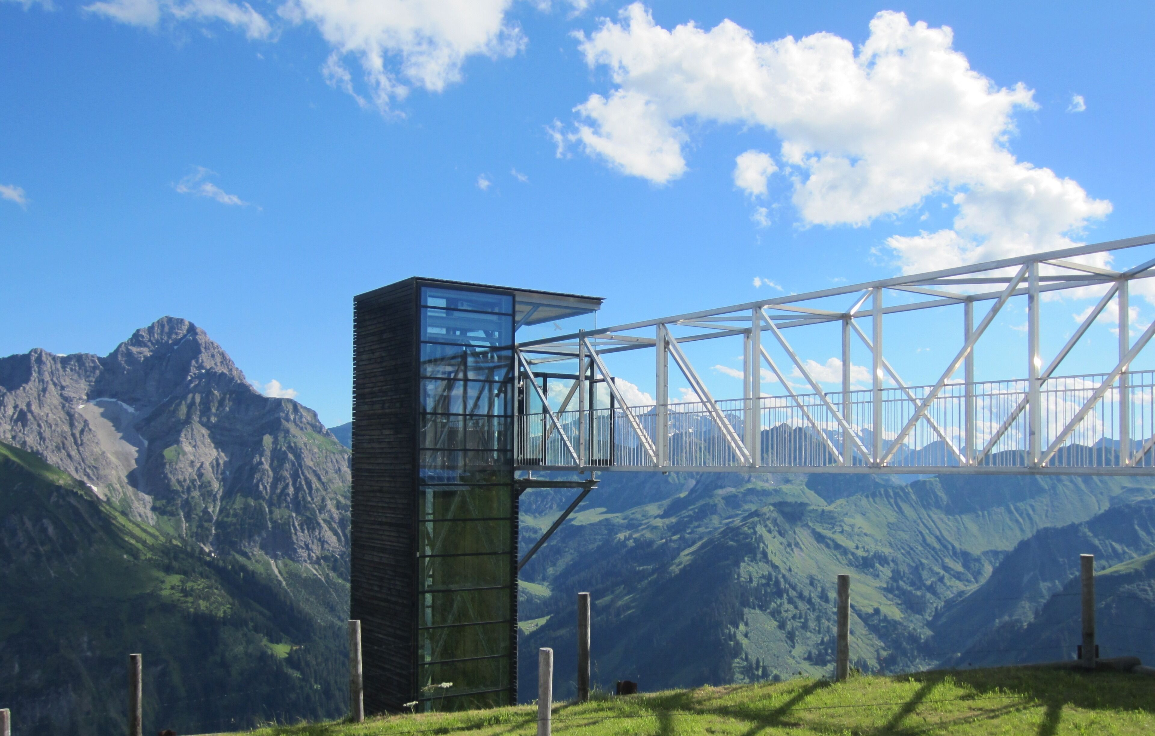 A modern glass building with a metal framework, set against a mountainous landscape and a clear blue sky.