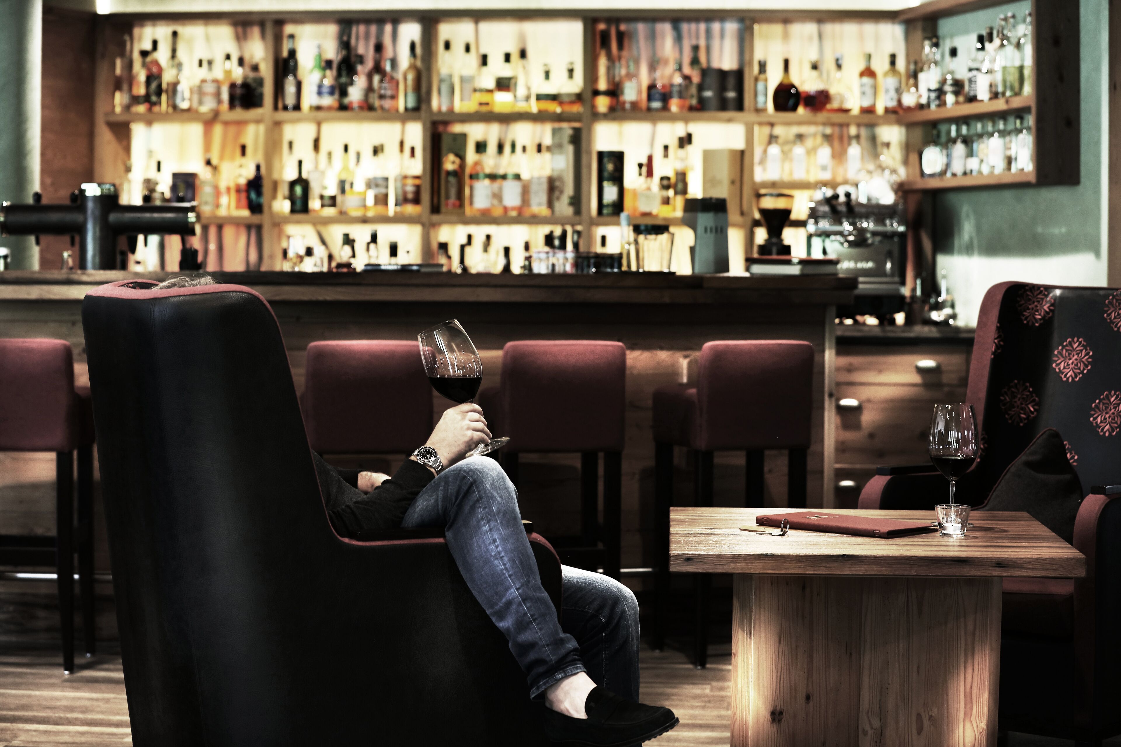 Un'area bar con un bancone in legno e scaffali pieni di bottiglie. Diverse poltrone imbottite e un tavolo con un bicchiere di vino.