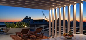 Une terrasse sur le toit, meublée de mobilier en bois, avec une table garnie de fruits et une vue sur le coucher de soleil au-dessus de la ville.