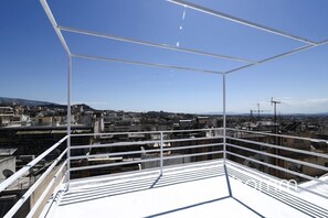 Une terrasse sur le toit, dotée d’une rambarde en métal, qui offre une vue sur un paysage urbain avec de nombreux bâtiments et des éoliennes visibles au loin.