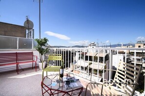 Un balcon avec un banc rouge, une chaise jaune, une table avec une bouteille et des verres, et un journal.