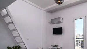 Une petite pièce avec un mur blanc, un sol carrelé noir, une chaise blanche, une petite table avec un bol, une échelle blanche, une plante en pot, un téléviseur à écran plat et un appareil de climatisation.