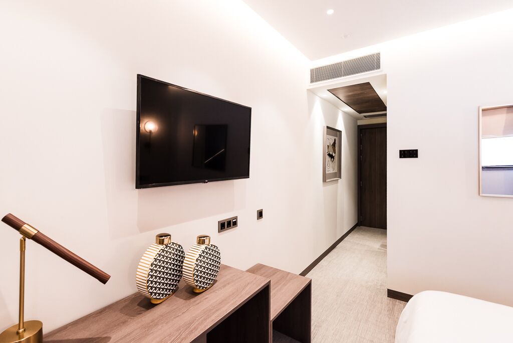 Um corredor moderno de hotel com uma TV de tela plana na parede, uma mesa consola de madeira com vasos decorativos e uma cama no quarto vizinho.