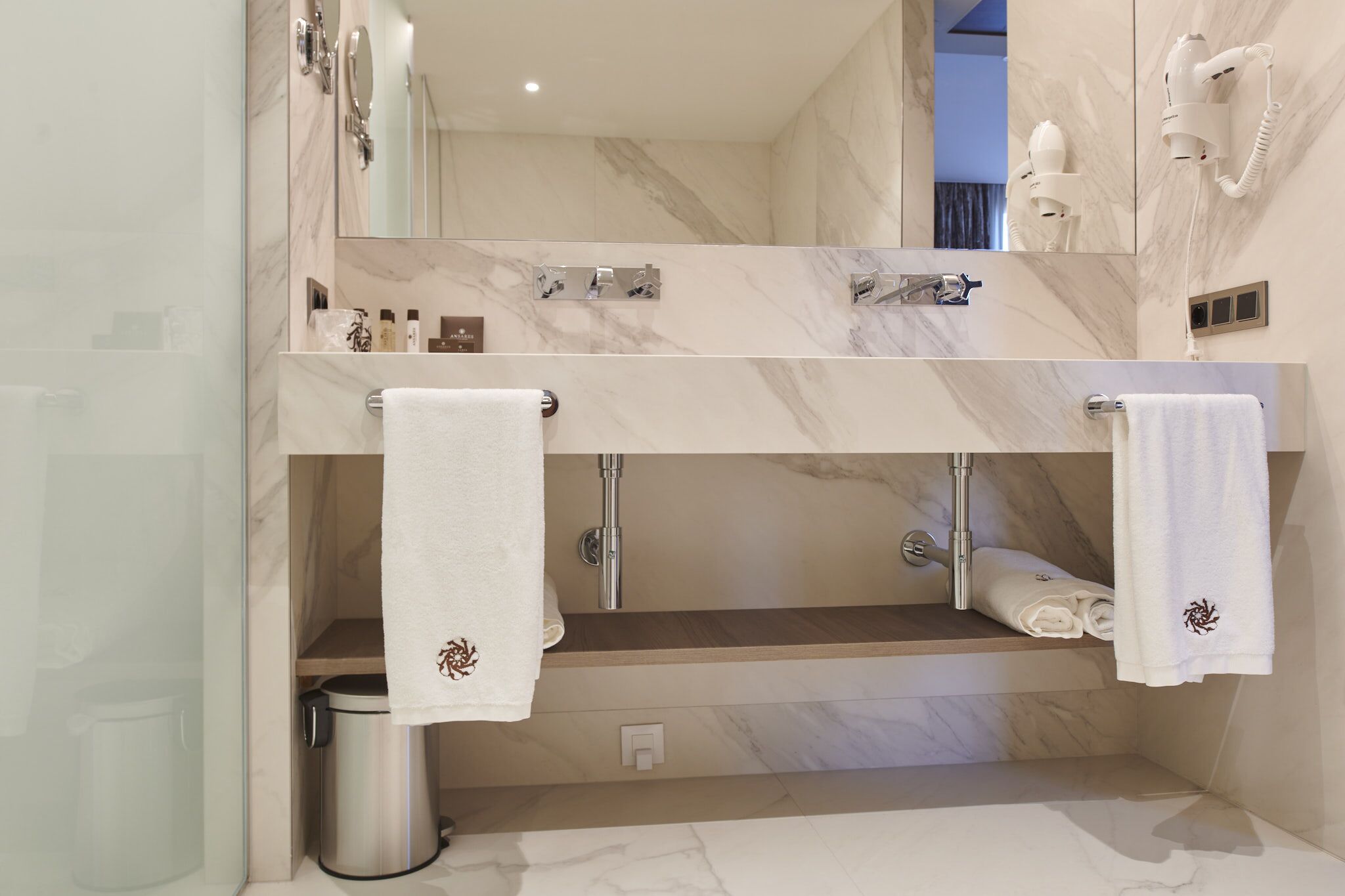 A modern bathroom with a double vanity, marble countertops, a large mirror, a towel rack, and a hairdryer mounted on the wall.