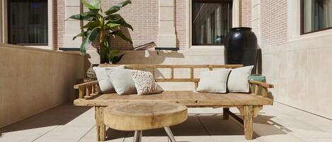 An outdoor seating area with a wooden bench and round table, surrounded by a brick wall and large windows.