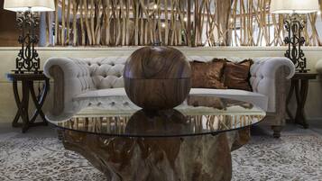 A room with a glass coffee table centered on a patterned carpet, flanked by a tufted sofa and two side tables with lamps.