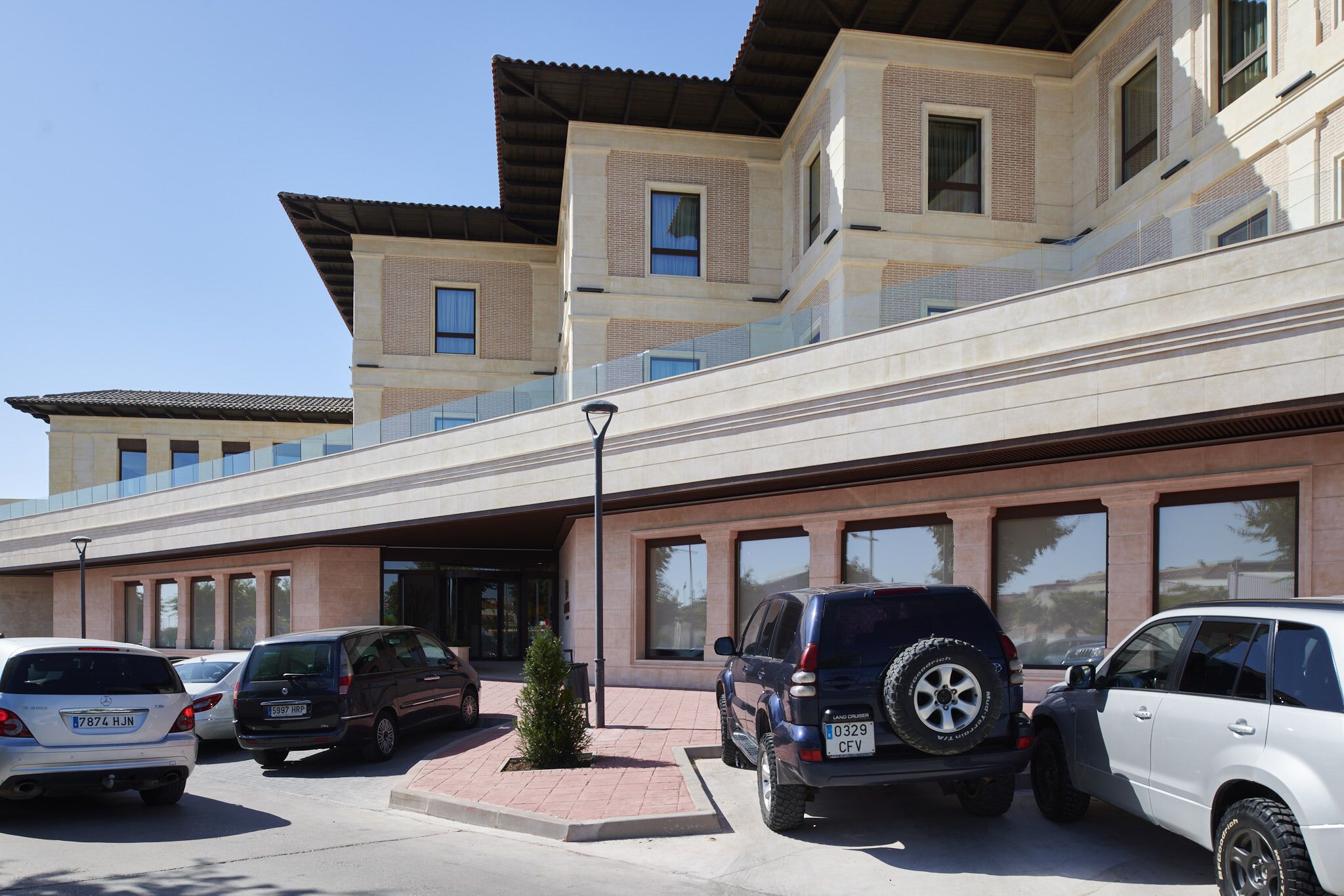 Ein modernes Gebäude mit Parkplatz. Mehrere Autos sind vor der Fassade geparkt, darunter ein schwarzer SUV und ein weißer SUV. Das Gebäude verfügt über große Fenster und ein flaches Dach.