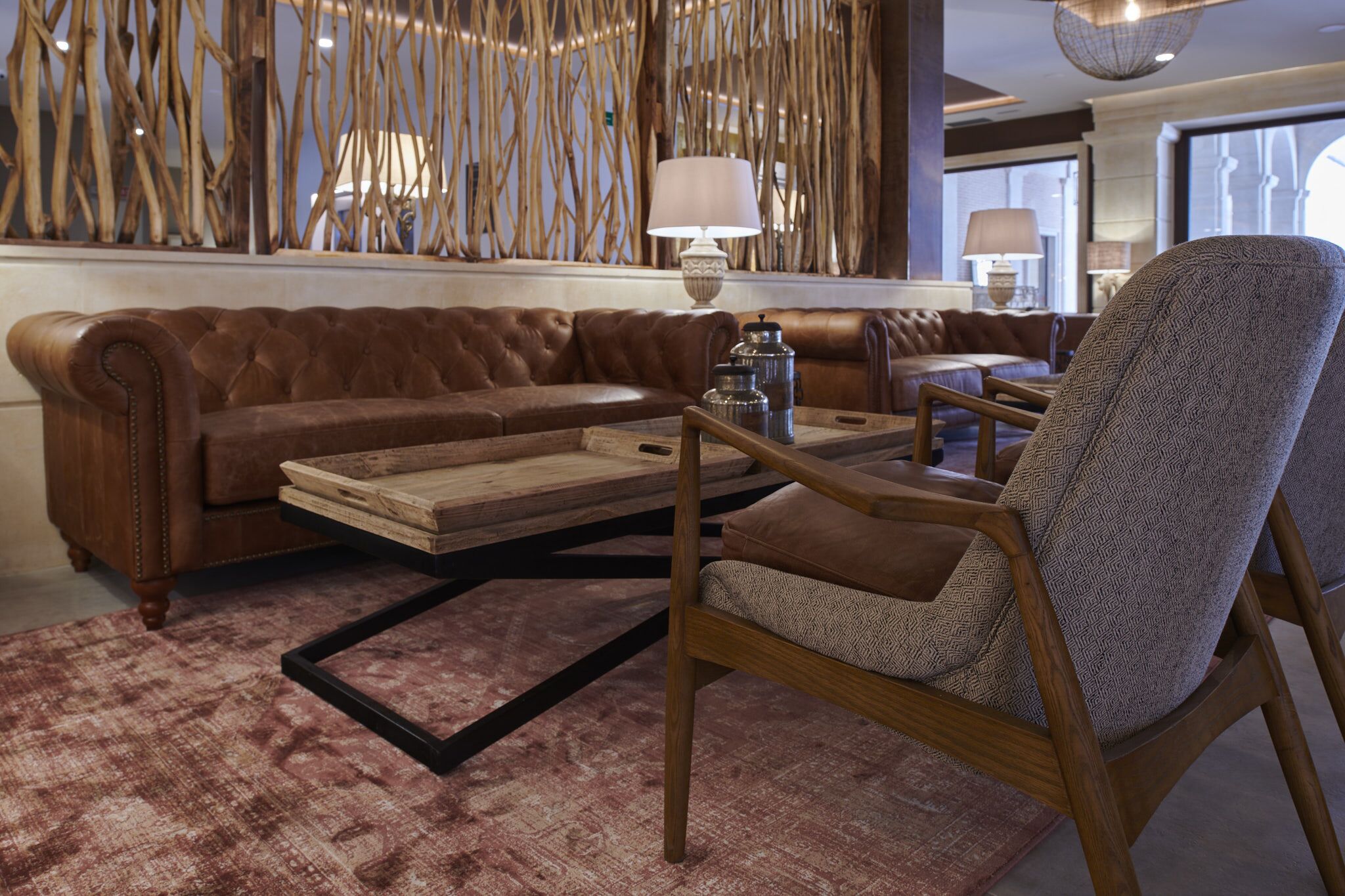 Un sofá de cuero marrón, un sillón estampado y una mesa de centro de madera en una sala de estar moderna con un muro decorativo y un ventanal grande.