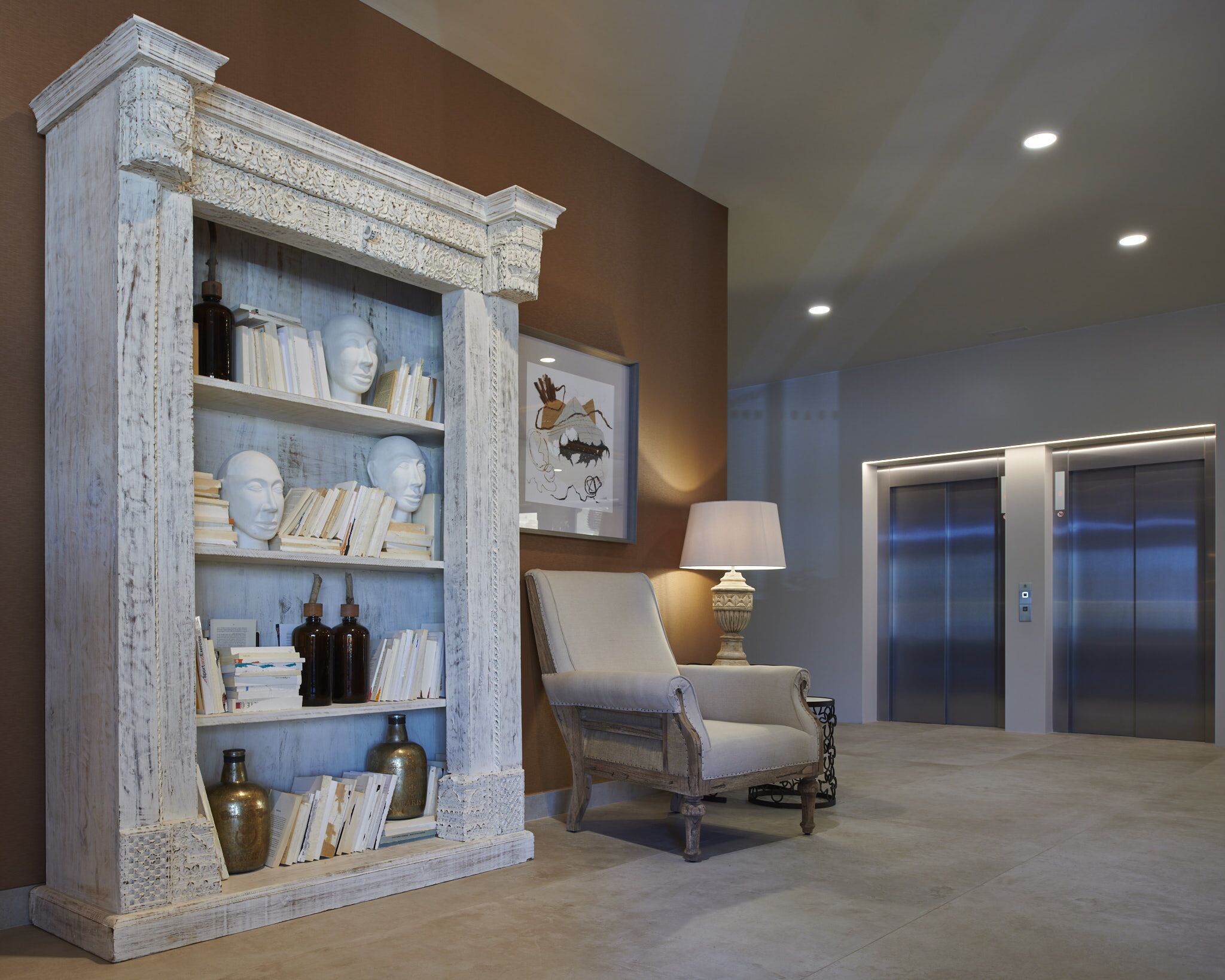 Un vestíbulo con una estantería blanca envejecida, un sillón blanco, una lámpara de pie y dos ascensores.