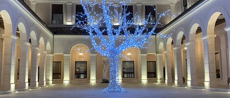 Un amplio patio con una escultura de árbol iluminada en el centro, rodeado por un edificio de estilo clásico con arcos y múltiples ventanas.