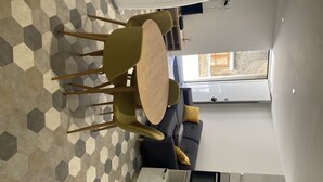 Ett modernt vardagsrum med ett hexagonalt mönstrat golv, en soffa med kuddar, ett runt träbord och en köksdel med en inbyggd ugn.