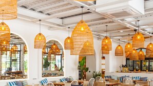 Een restaurant met houten tafels en stoelen, hanglampen van riet en grote ramen.