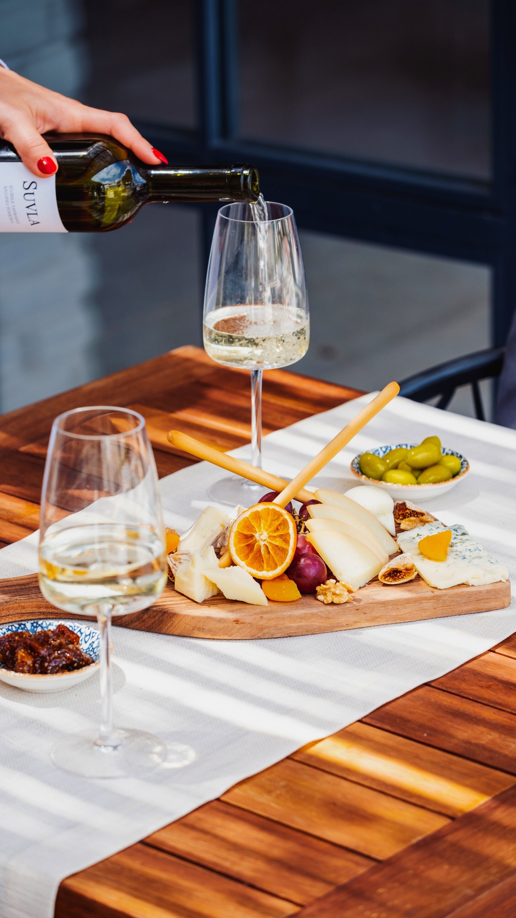 A wooden table with a charcuterie board, two glasses of white wine, and a bottle being poured into one of the glasses.